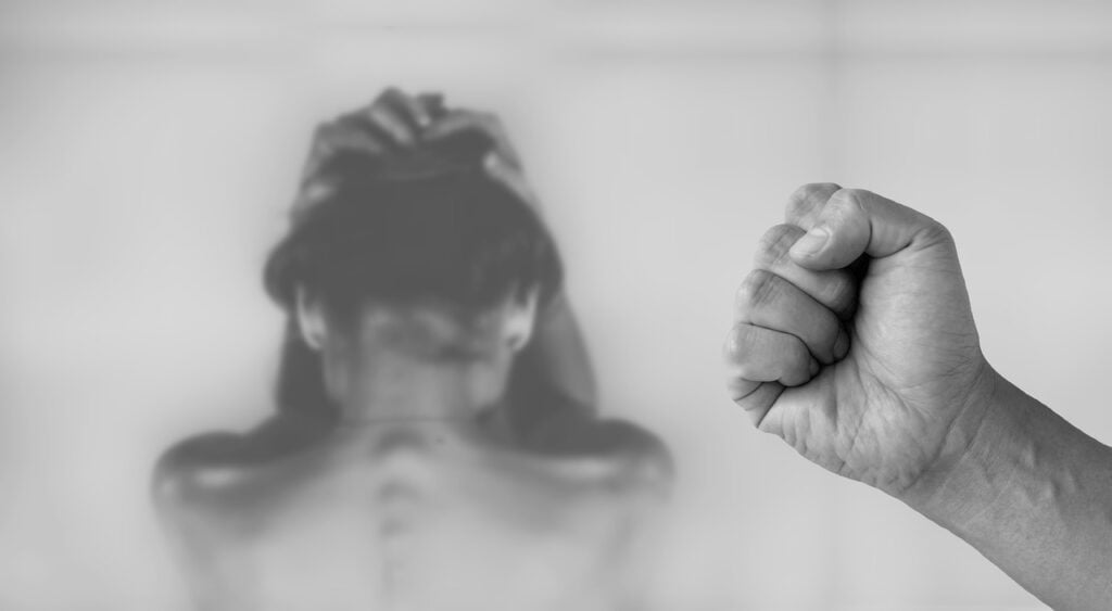 Das Bild steht symbolisch für das Thema des Artikels: Ethnosexismus. Das Foto ist in schwarzweiß. Man sieht darauf eine Frau mit dem Rücken zum Betrachter, die ihre Hände zu Schutz über ihren Kopf zusammenhält. Im Vordergrund des Bildes sieht man ein männliche zu Faust geballten Hand.