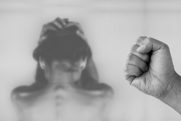 Das Bild steht symbolisch für das Thema des Artikels: Ethnosexismus. Das Foto ist in schwarzweiß. Man sieht darauf eine Frau mit dem Rücken zum Betrachter, die ihre Hände zu Schutz über ihren Kopf zusammenhält. Im Vordergrund des Bildes sieht man ein männliche zu Faust geballten Hand.