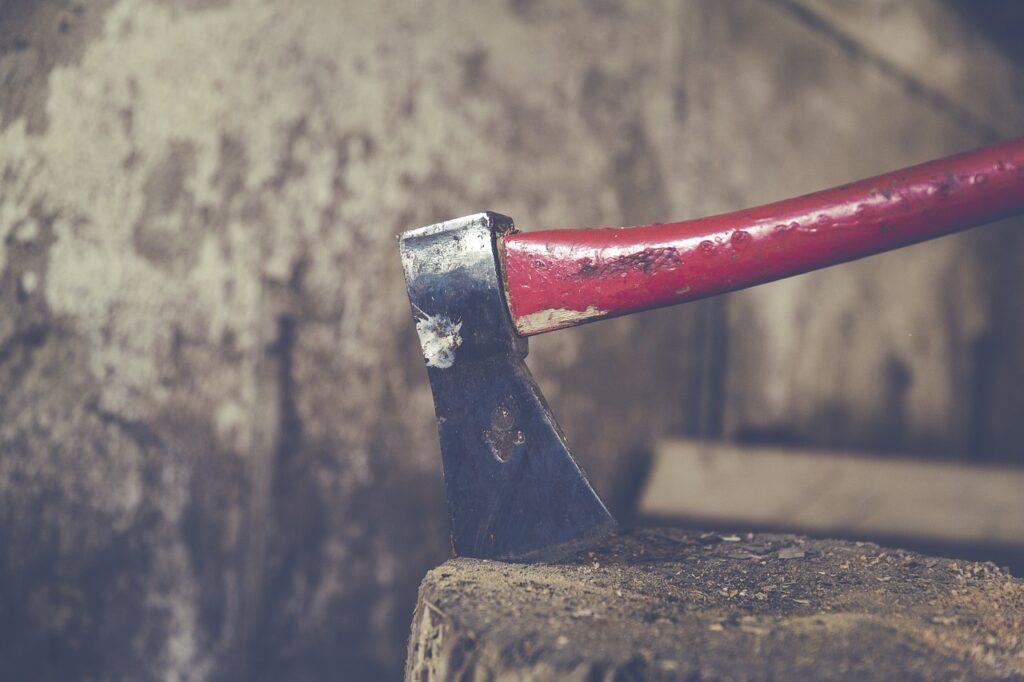 Beitragsbild: Penis abgeschnitten – Frau attackiert ihren Partner im Schlaf. Das Foto zeigt eine Axt mit rotem Stil, die einem Holzblock steckt. Dahinter ist eine alte, abgenutzte Mauer zu sehen.