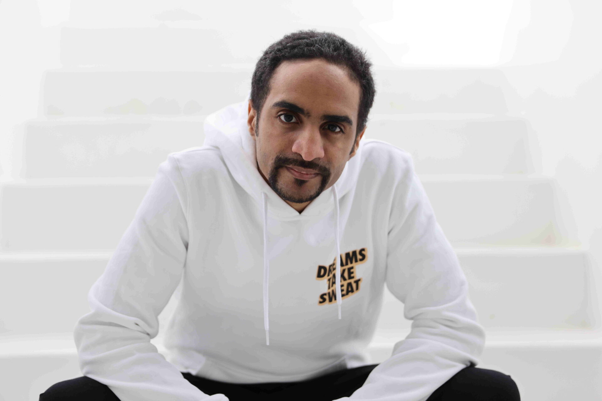 Auf dem Foto sieht man den Wiener Rapper AYE-sight. Er trägt einen weißen Pullover und eine schwarze Hose, während er hockt und direkt in die Kamera blickt. 