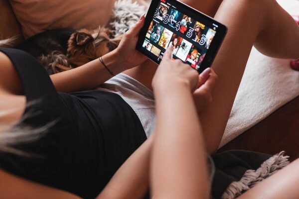 Auf dem Foto sieht man eine Frau sitzend auf einer Couch. Das Foto ist dabei von einem Winkel aufgenommen rechts schräg über ihren Kopf. Das Sofa ist Lachsfarben und man erkennt ein großes Sofakissen links neben ihr. Rechts von sieht man eine Hand, die auf das Tablet zeigt, das die erste Frau in der Hand hält. Man sieht das Netflix Menü auf dem Tablet anscheinend zeigt die zweite Frau auf eine Netflix Serien Empfehlung