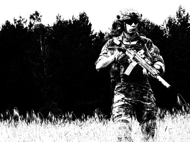 EU-Aufrüstung: Das Bild zeigt einen Soldaten in voller Kampfmontur, der mit einem Sturmgewehr bewaffnet ist. Er trägt einen taktischen Helm mit Schutzbrille und ein Tarnmuster-Uniform. Der Soldat steht in einem Feld mit hohem Gras, und im Hintergrund ist eine dichte Baumreihe zu sehen. Die Aufnahme hat einen starken Schwarz-Weiß-Kontrast, wodurch das Bild einen stilisierten, fast grafischen Look erhält. Die Haltung des Soldaten wirkt aufmerksam und kampfbereit.