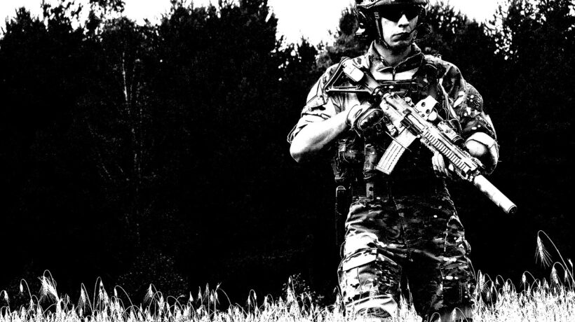 EU-Aufrüstung: Das Bild zeigt einen Soldaten in voller Kampfmontur, der mit einem Sturmgewehr bewaffnet ist. Er trägt einen taktischen Helm mit Schutzbrille und ein Tarnmuster-Uniform. Der Soldat steht in einem Feld mit hohem Gras, und im Hintergrund ist eine dichte Baumreihe zu sehen. Die Aufnahme hat einen starken Schwarz-Weiß-Kontrast, wodurch das Bild einen stilisierten, fast grafischen Look erhält. Die Haltung des Soldaten wirkt aufmerksam und kampfbereit.